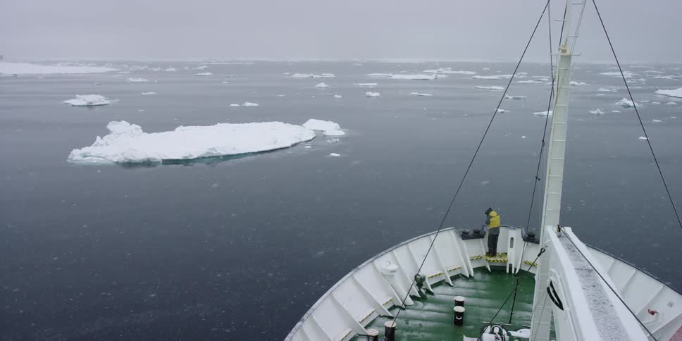 A man stands at the front of a boat on icy waters