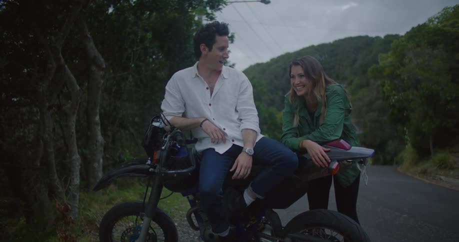 A man and woman laugh while leaning on a motorcycle