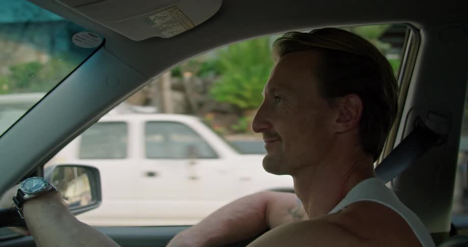 Blonde man with white undershirt and tattoo drives car in the street passing by vehicles smiling