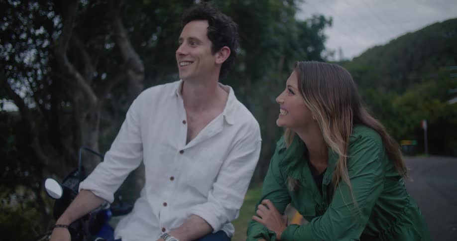 A man and woman are outside talking and smiling while relaxing near green hills and trees