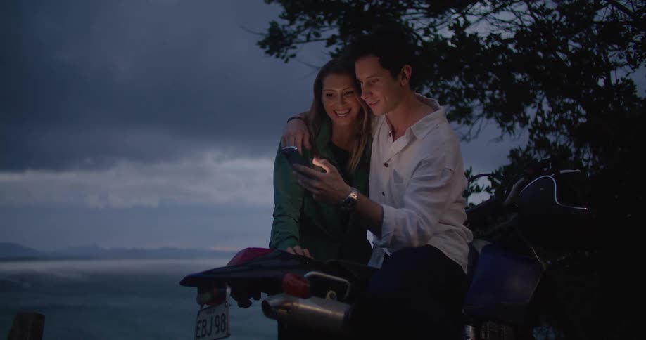 A man sitting on a motorcycle with his arm around a woman scrolls on his phone as they talk and smile