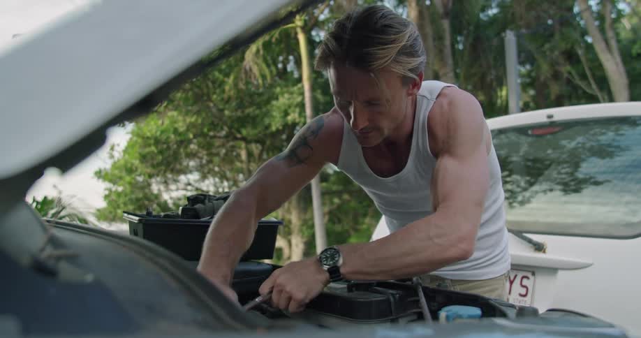 A mans arm muscles bulge as he uses a socket wrench while repairing a car outside