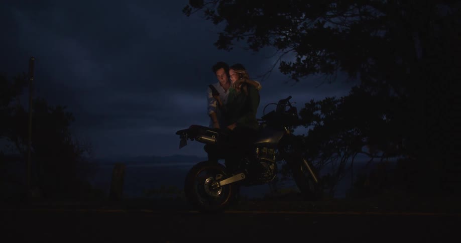 A man and woman standing close to each other and looking at a smartphone next to a motorcycle at night