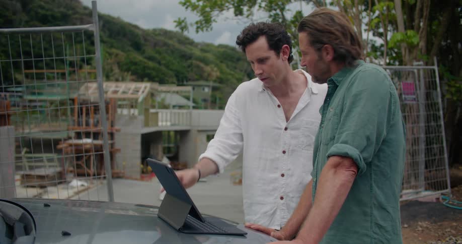 Two men are leaning against a car looking at a laptop and waving and talking