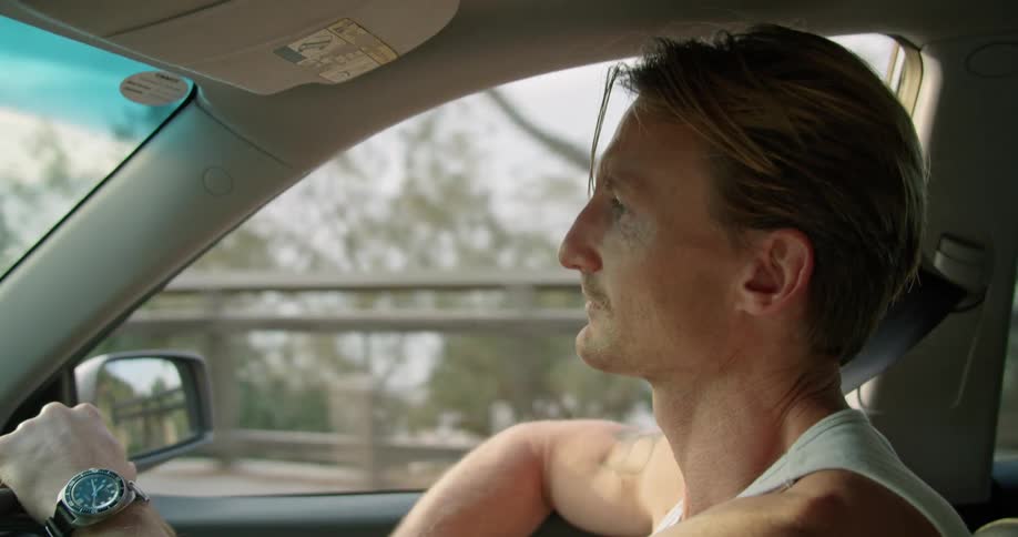 Blonde man with white undershirt and tattoo drives car next to shore crossing the bridge