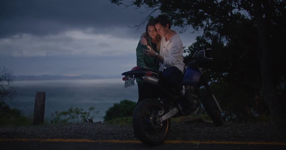 A man is sitting on a motorcycle next to a woman and scrolling on a cell phone and smiling