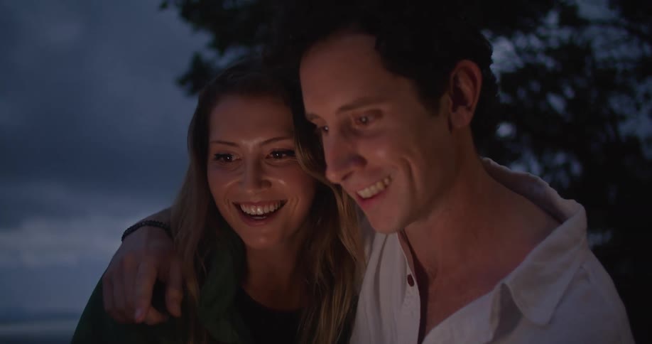 Man and woman sit near sea at night hugging and laughing while looking at mobile phone