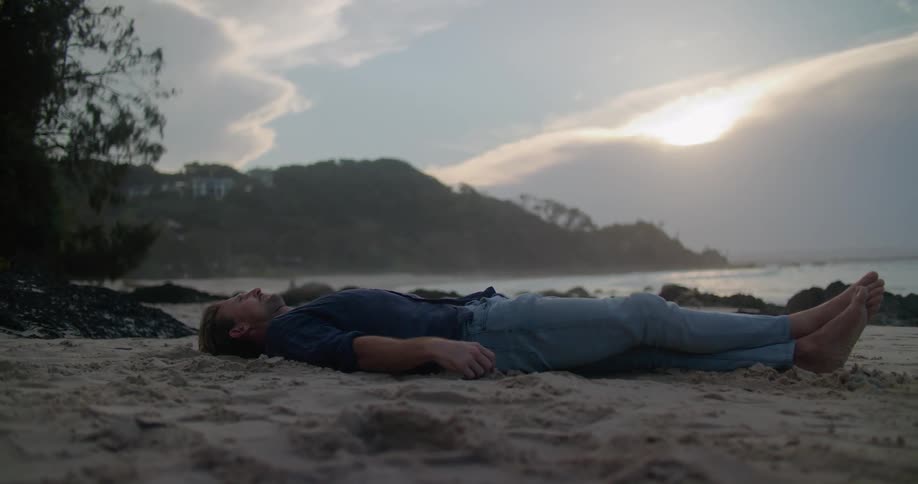 A man is lying barefoot on a beach as the sun shines from behind a cloud