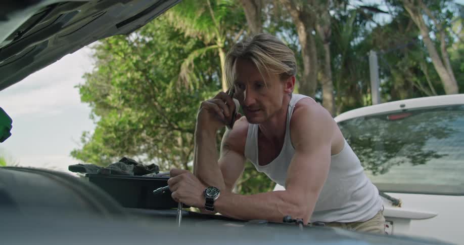 A man is talking on a cellphone and then continues to work under the hood of a car outside during the day