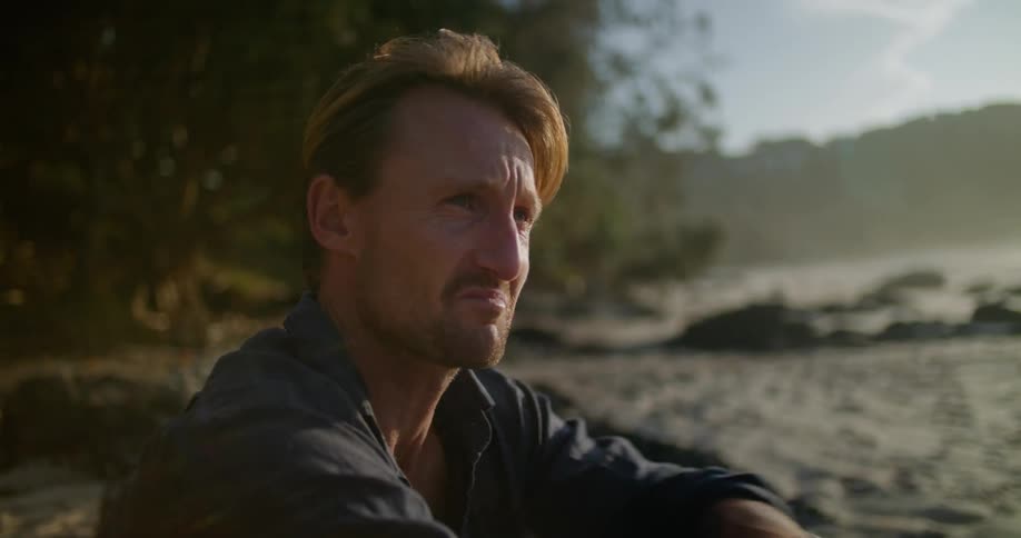 Blonde man with blue shirt sitting on beach and looks at sea crying