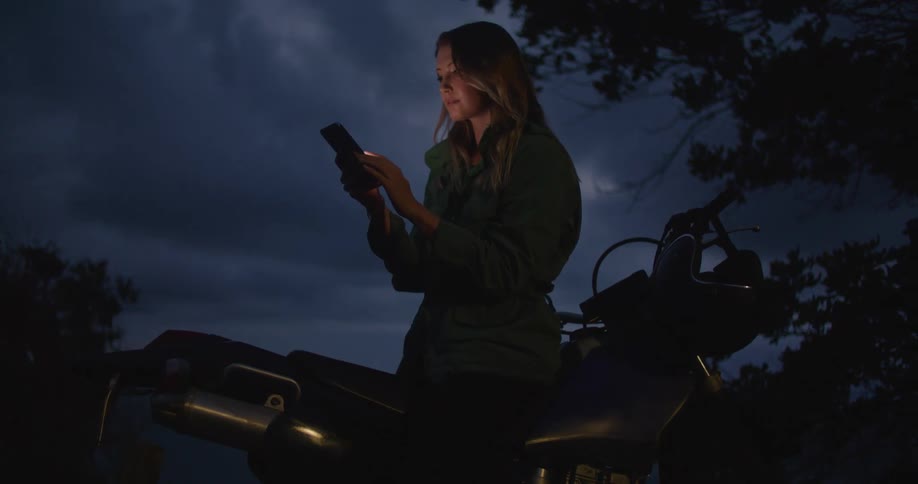A woman is interacting with a smartphone while outdoors straddling a motorcycle at night
