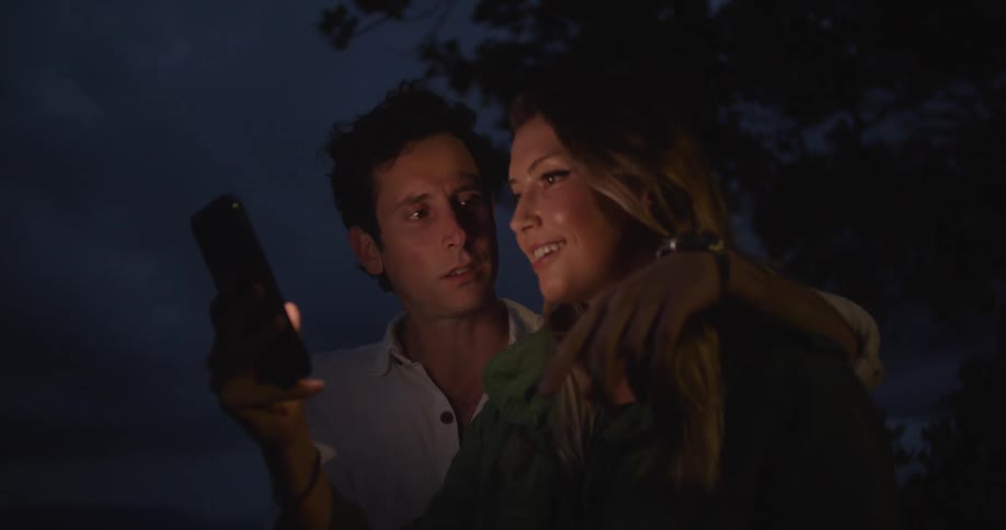 A man and a woman are both looking at a smartphone and having a conversation while standing outdoors at night