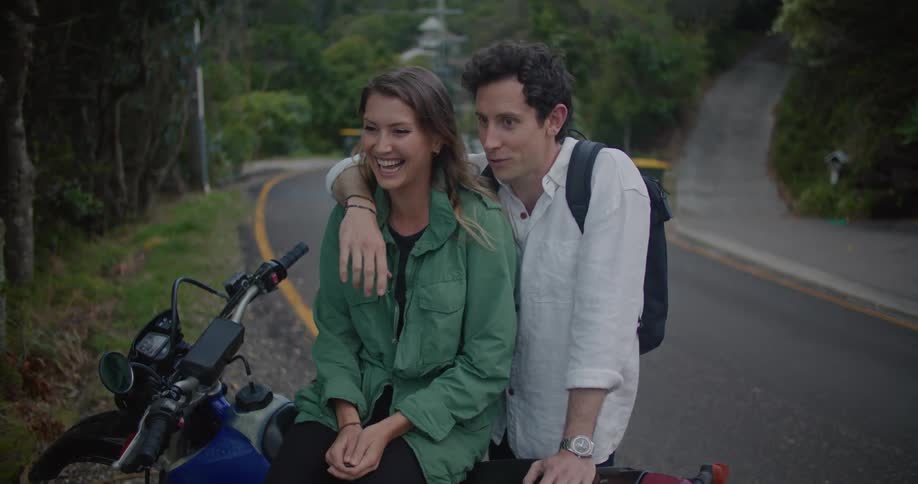 A man stands with his arm around a woman sitting on a motorcycle as they talk and smile