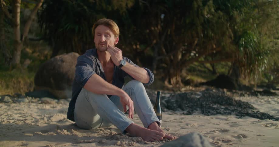 A man is sitting on a beach next to a wine bottle staring off and rubbing his face