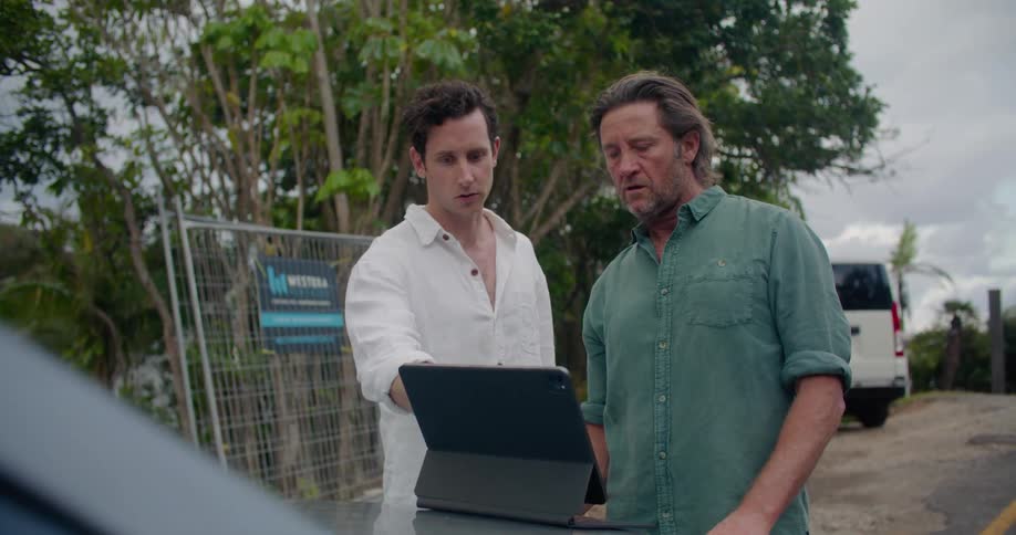 Men with white and green shirts discuss matter looking at tablet on car hood on the road side