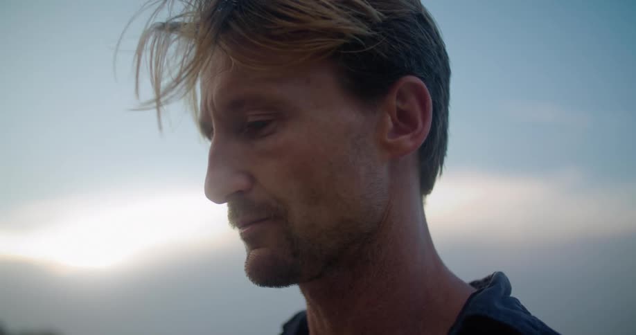 A man is standing outside on a windy sunset evening and drinking from a wine bottle