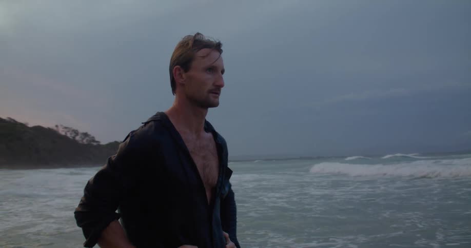 A fully clothed man stands in the surf bracing himself as waves splash