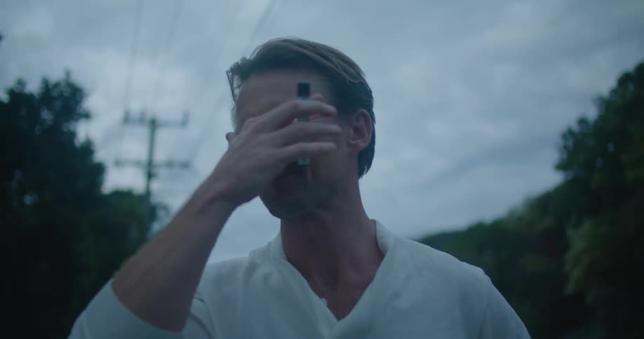 A man is walking along a mountain road at dusk while smoking an electronic cigarette when he stops and stares into the camera