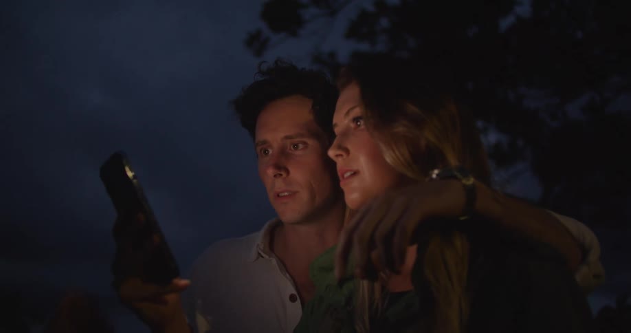 A man is hugging a woman while they both are interacting with a smartphone while standing outdoors at night