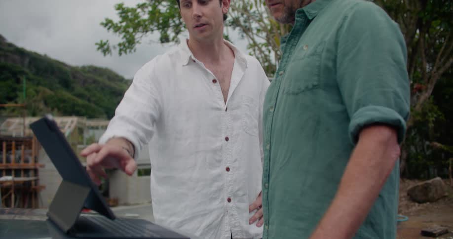 Two men are outside looking at a laptop and talking at a building construction site