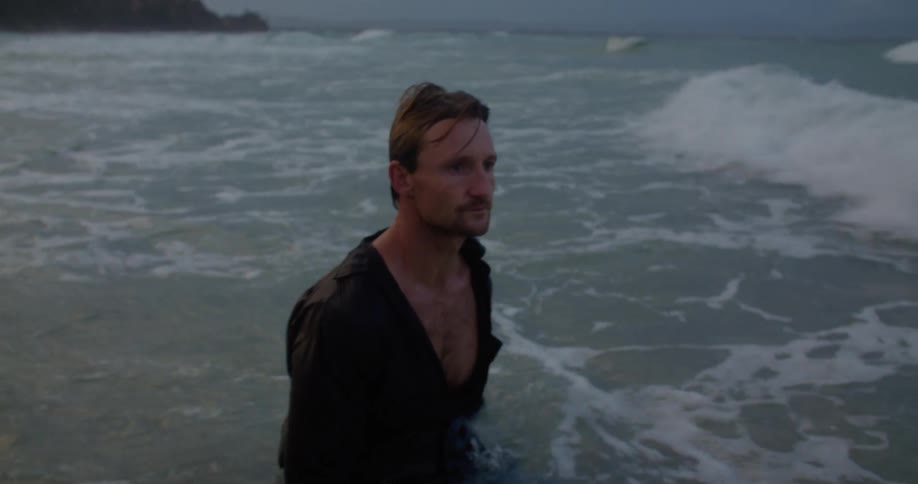 Man wearing jeans and shirt lays down in water near beach and lets waves roll over him