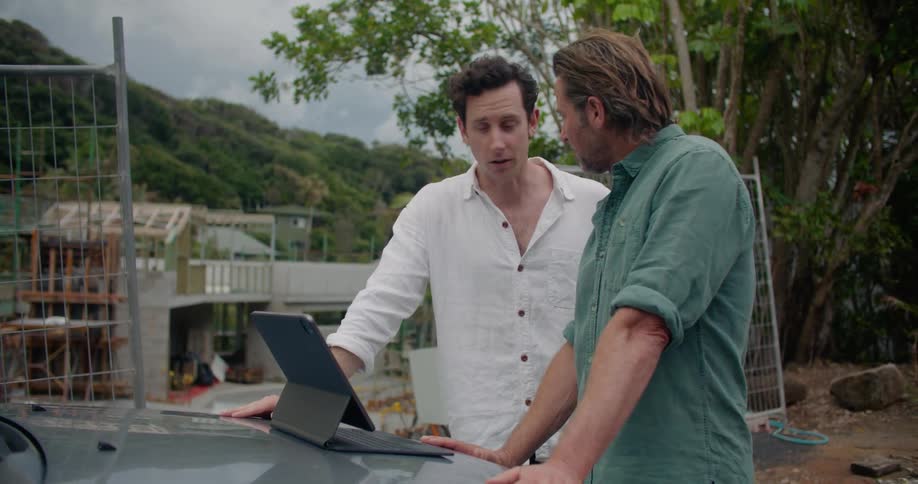 Two men look at a laptop and talk on a construction site