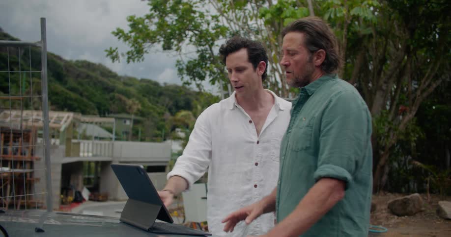 Two men stand near car parked at construction site discussing tablet contents with forest covered mountain in background