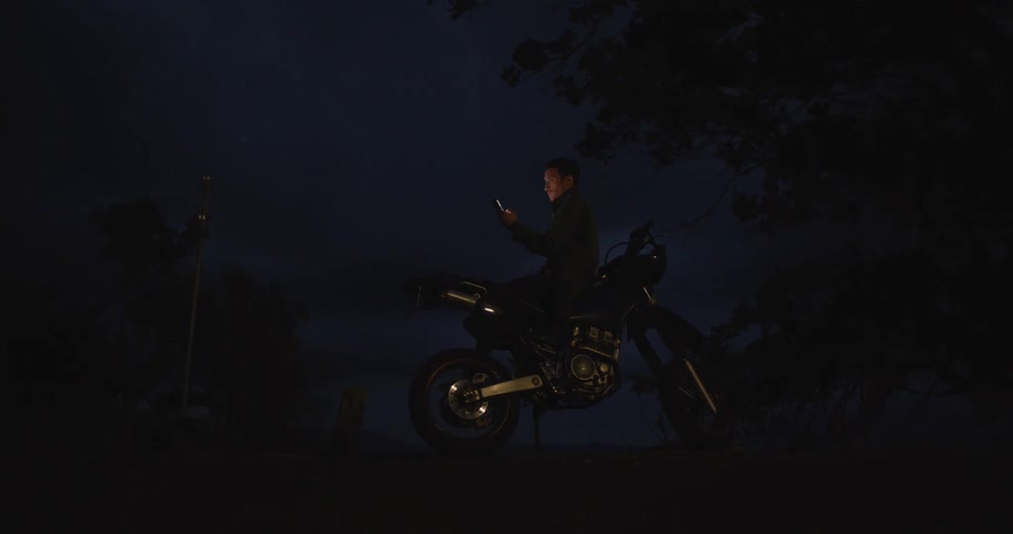 Man reclined on a parked motorcycle checking his smartphone at night