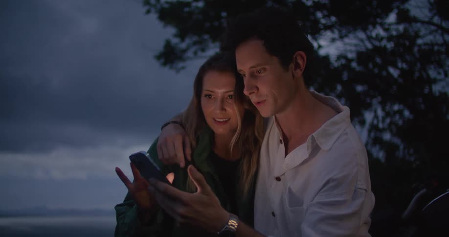 A man and woman are standing outside at night scrolling on a cell phone and smiling