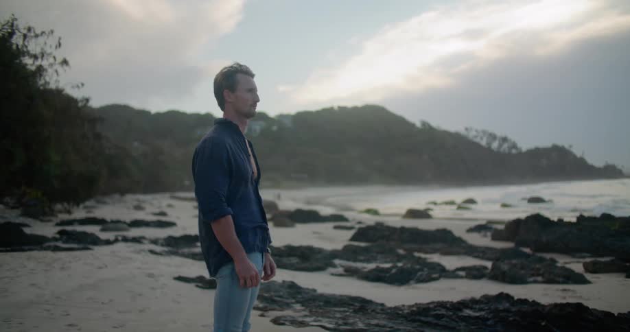 A man standing on a beach looking out at the ocean as the wind blows his shirt