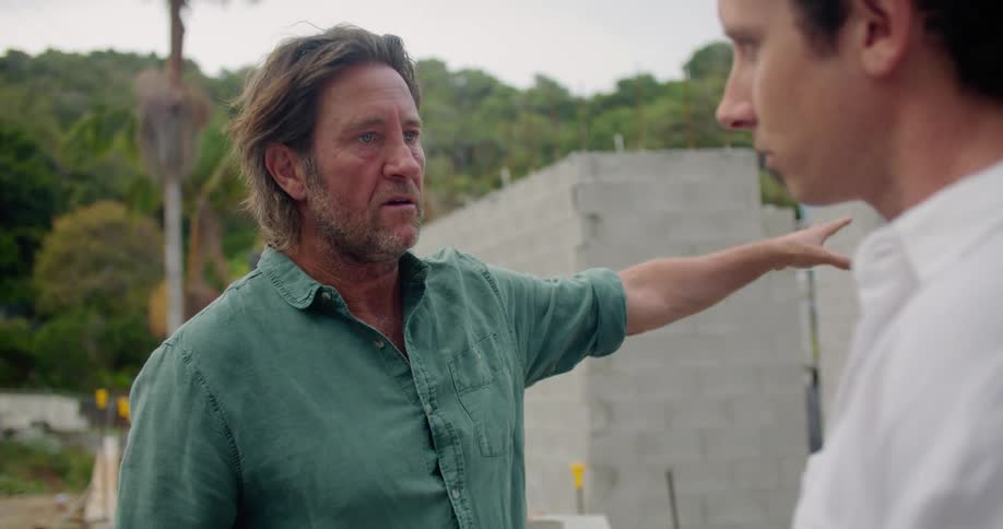 Two men are discussing building plans on a residential construction site in a forested area during the day