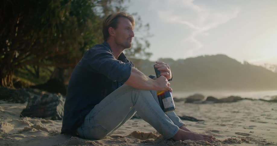 Young barefoot man sitting on the sand on a beach nex to the ocean drinking wine from a bottle at sunset