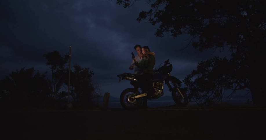 Man and woman sitting on motorbike use smartphone outdoors at night