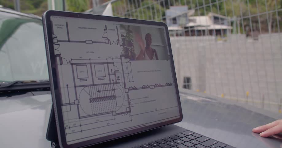 A man waves at a computer screen with a woman in the corner next to blueprints as they video chat
