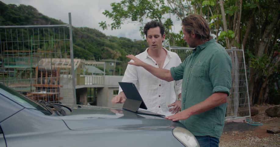 Two men prop a laptop on the back of a vehicle while at a construction site