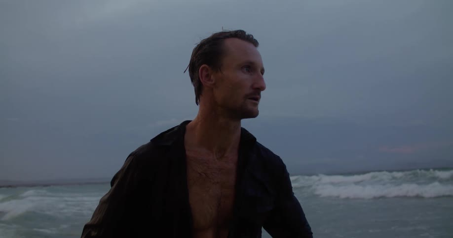 A man standing on the beach with wet hair and shirt as the waves roll in behind him