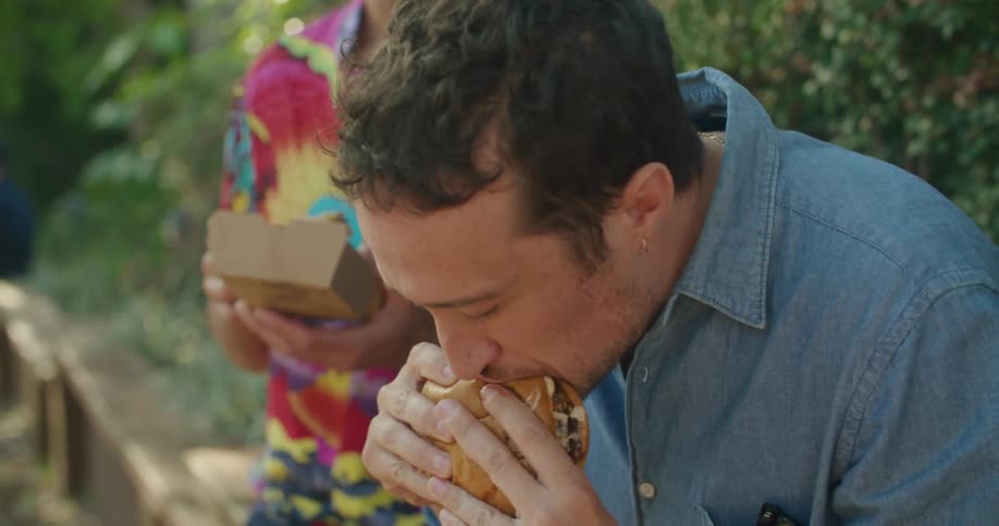 Young male couple eating burgers with french fries on a bench outdoors