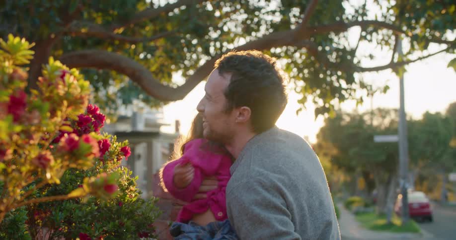 Father and daughter smile as he picks her up near flower beds on sunny day