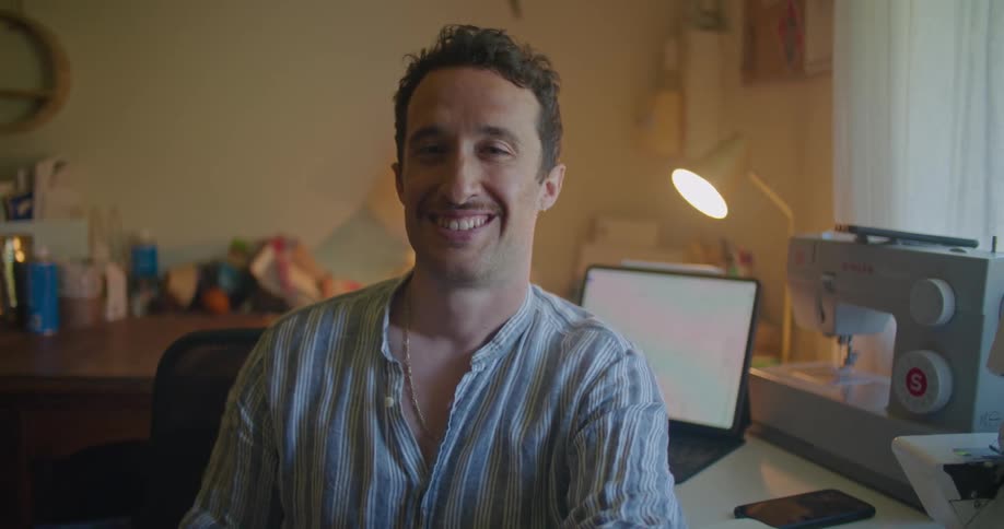 Man sits and laughs with back to table holding tablet and sewing machine