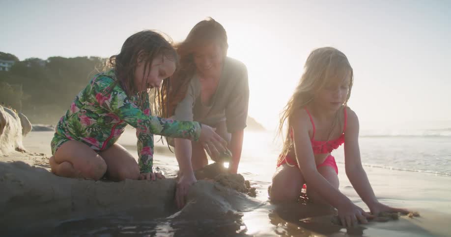 A woman knees on the beach and piles sand up with her two young daughters as a man walks past