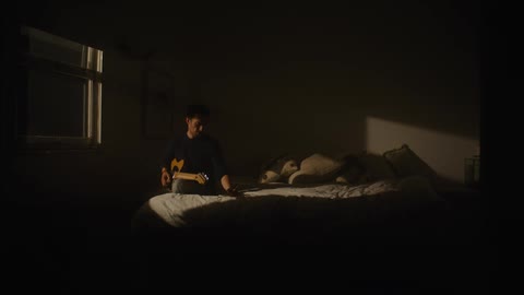 Man with black sweater and jeans plays guitar on bed in dark room at night