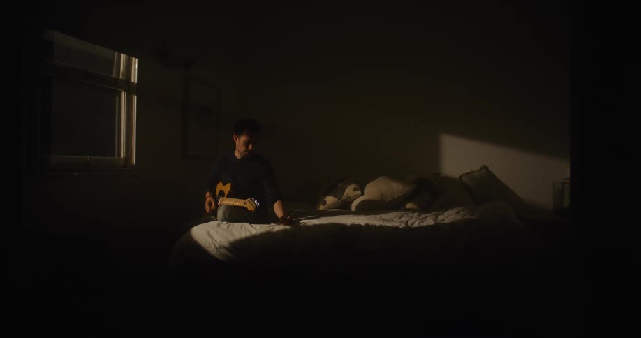 Man with black sweater and jeans plays guitar on bed in dark room at night