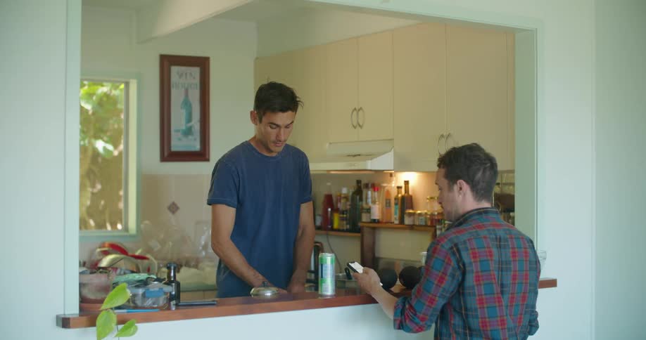 Man sitting in the kitchen at home shows his phone to a friend cooking