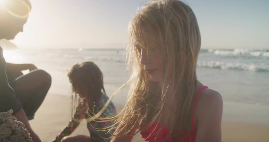 Girl in red swimsuit playing with sand on the beach near her sister and father at sunset
