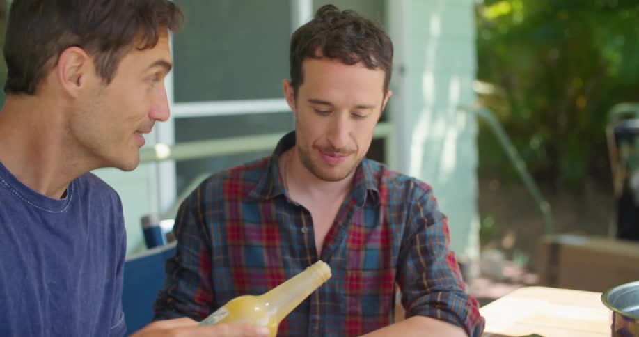 Two men pour sauce on bowls of food as they eat on a picnic table outside
