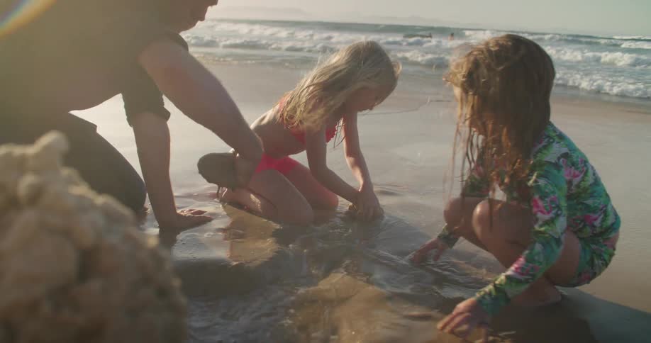 Father and daughters playing with sand on the beach next to the ocean at sunset