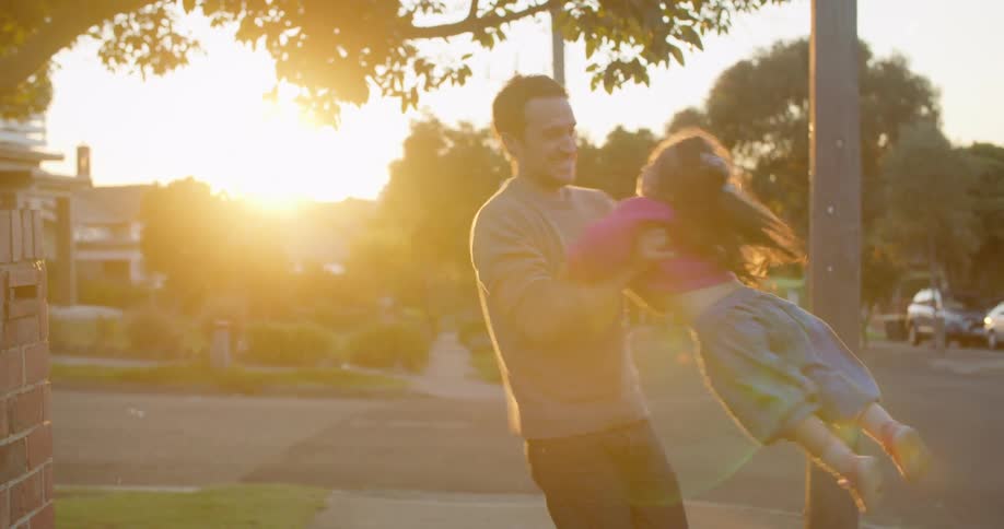 A father is swinging his daughter around in circles on a neighborhood sidewalk at sunset