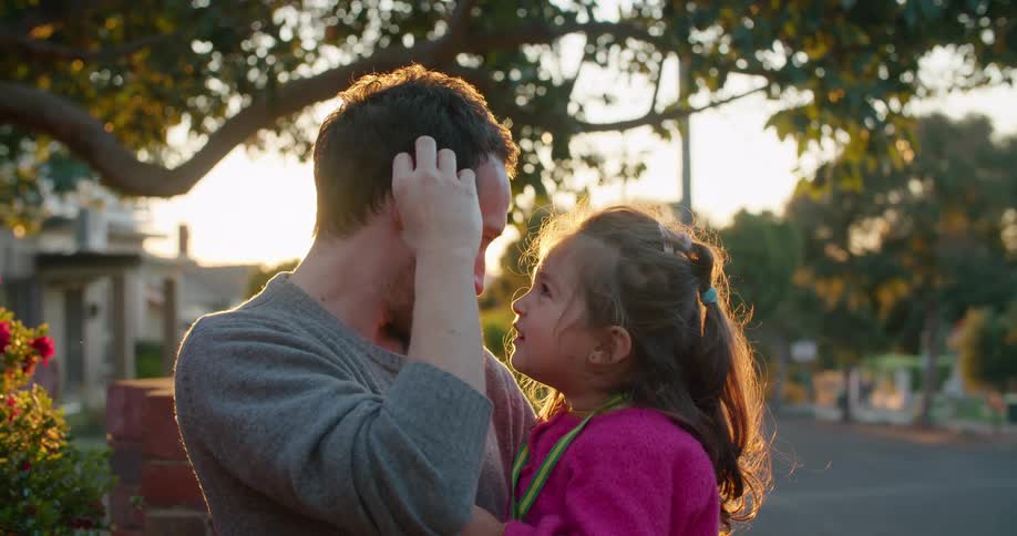 A dad picks a flower from a bush and places behind his ear causing his daughter to laugh