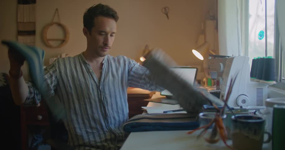 A man sits at a table with a sewingmachine and a computer and looks at his cell phone and then starts flipping through material