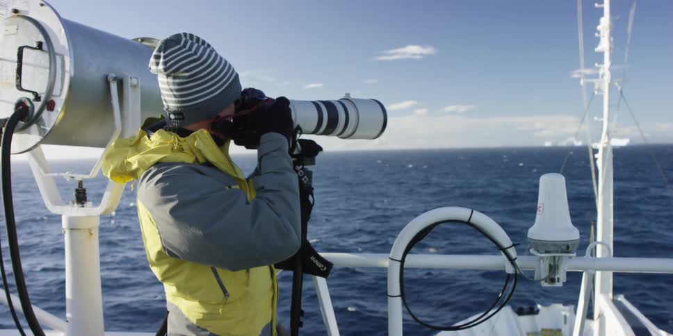 A photographer uses a long zoom lens camera while standing on a boat
